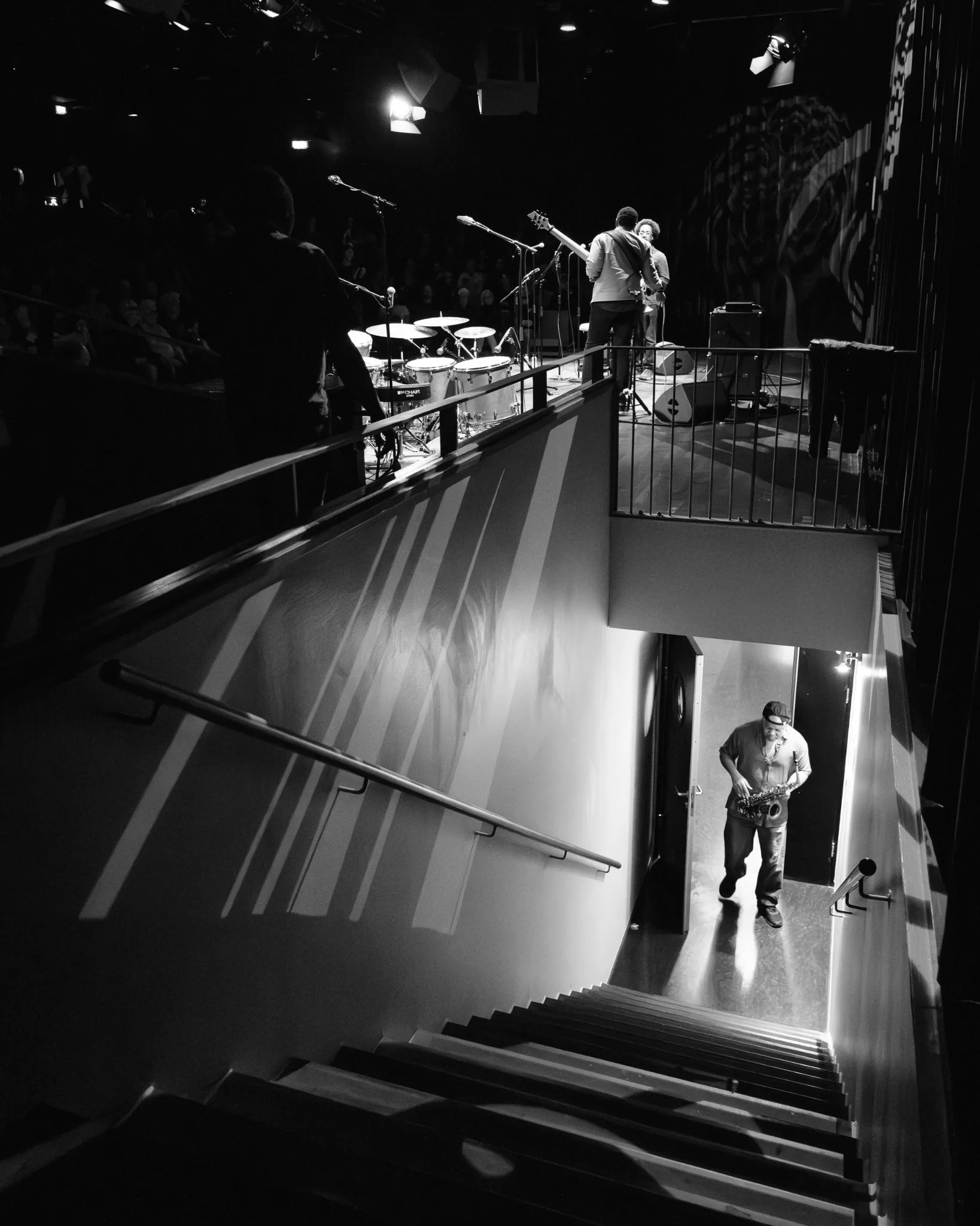 Black-and-white view of a musician on stage above a lit corridor with two people.
