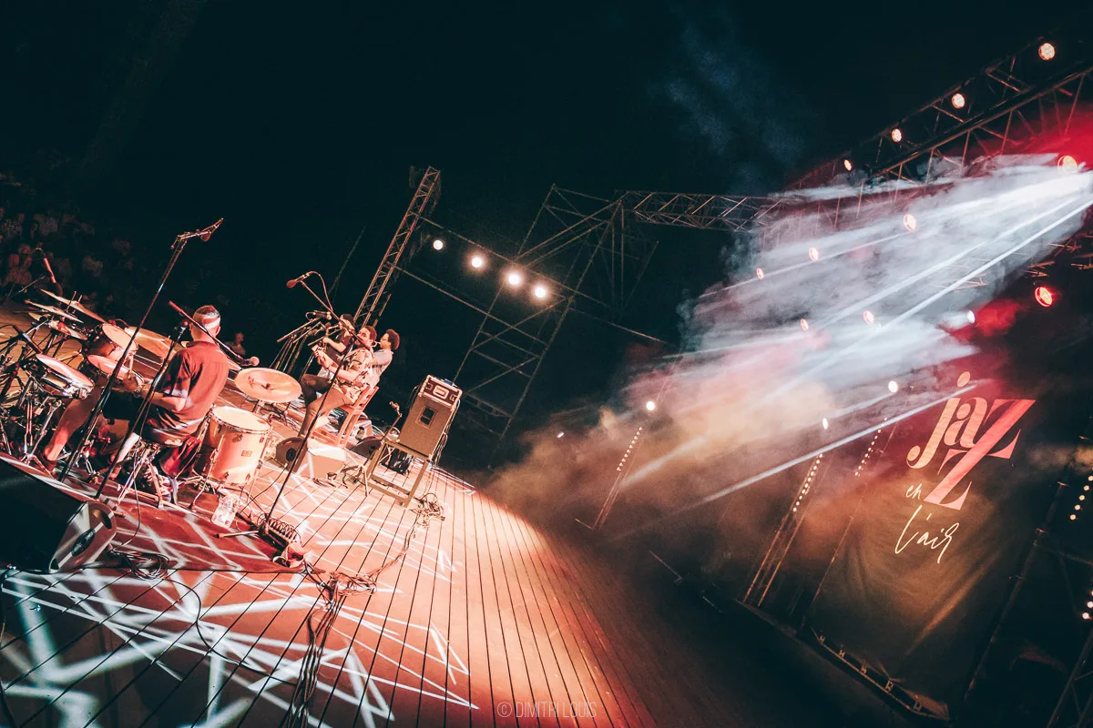 Angled view across the stage, musicians and microphones beneath white beams of light.