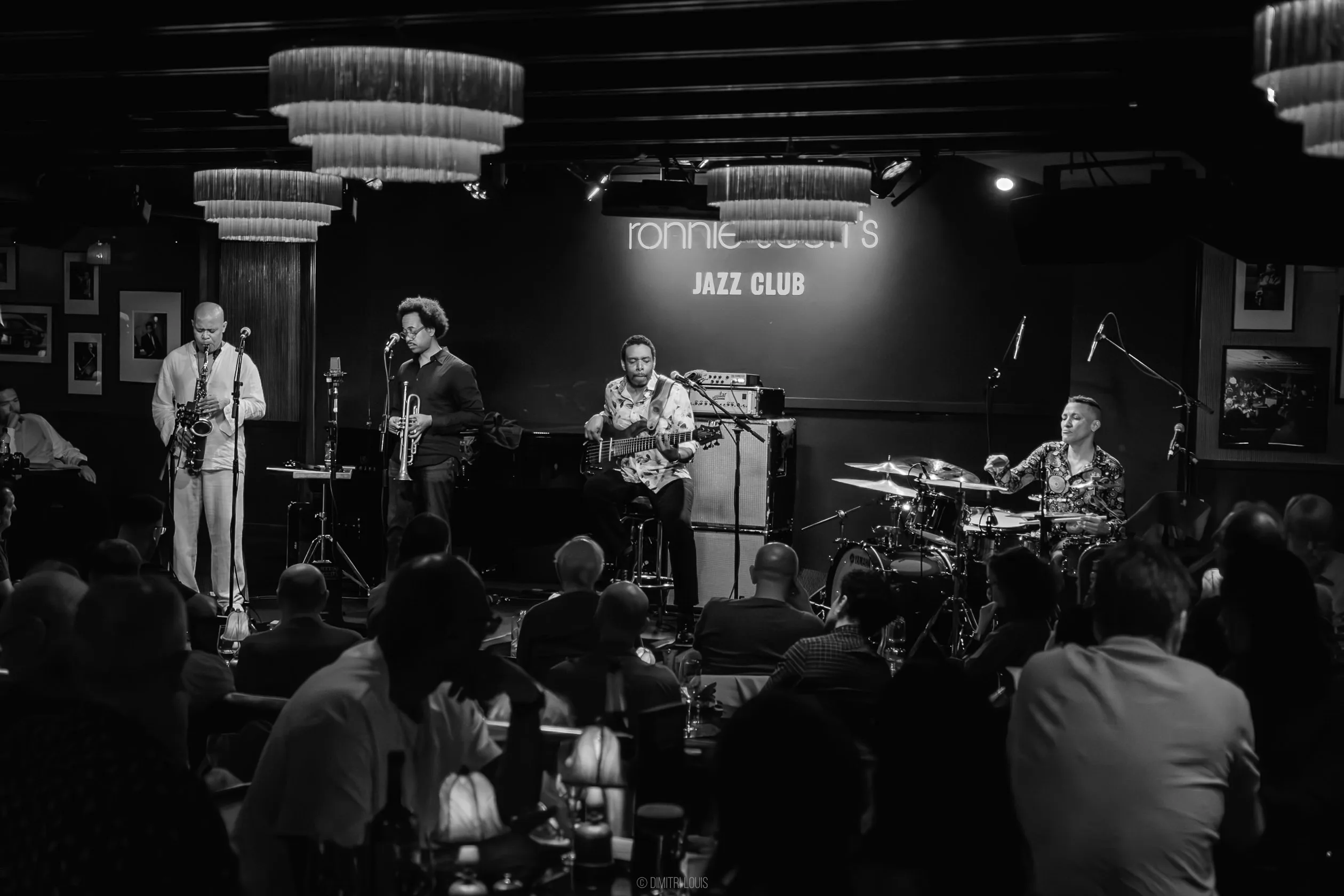 Black-and-white view of the quartet on stage, with saxophone and trumpet at left, electric bass in the centre and drums at right.