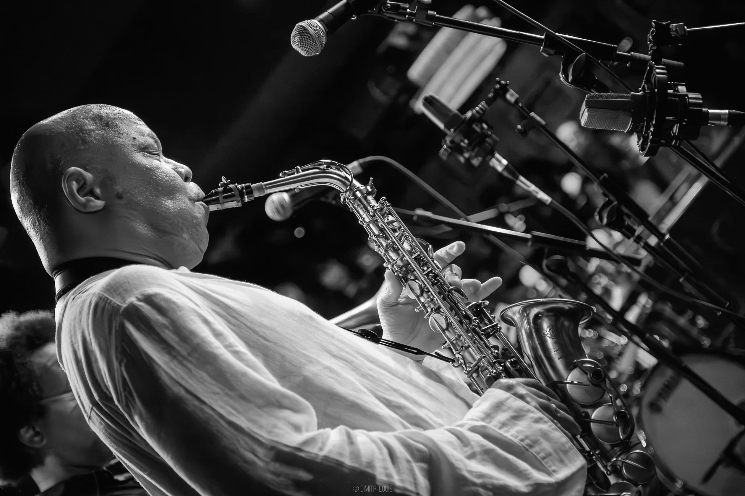 Black-and-white close-up of Steve Coleman playing alto saxophone, photographed from below with microphones overhead.