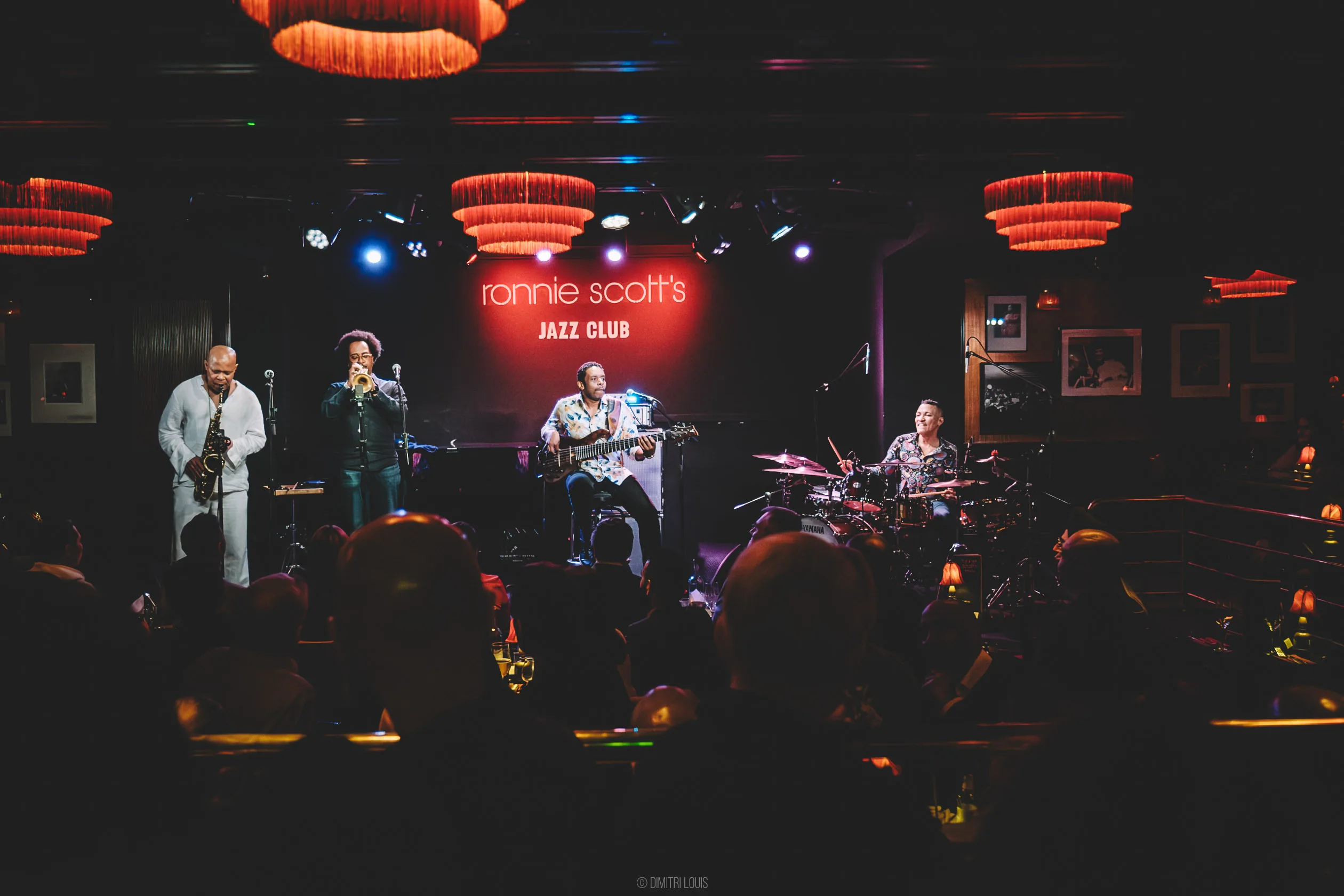 Colour view across audience tables towards Five Elements, with Jonathan Finlayson playing trumpet beneath the Ronnie Scott’s sign.